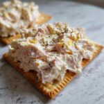 A close-up of creamy ham salad recipe piled high on a square cracker, garnished with dried herbs.