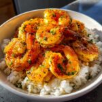 Close-up of juicy Hawaiian garlic shrimp coated in a rich, orange sauce and sprinkled with parsley, served over white rice in a bowl.