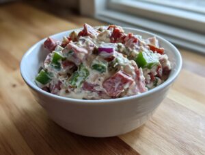 A white bowl filled with creamy, chunky hoagie dip featuring diced meats, green peppers, and red onion.