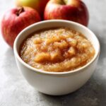 A close-up of chunky homemade applesauce topped with cinnamon in a white bowl, with fresh red apples in the background.