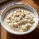 A close-up of a white bowl filled with rich, creamy cream of chicken soup, topped with shredded chicken and herbs.