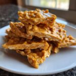 A stack of golden brown, crunchy pieces of homemade peanut brittle recipe piled on a white plate.
