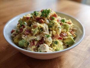 A close-up of a white bowl filled with creamy honey mustard chicken salad mixed with chunks of avocado and crispy bacon pieces.