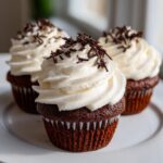 Three rich chocolate hot chocolate cupcakes topped with swirls of white frosting and chocolate shavings.