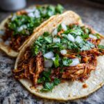 Two delicious chicken tacos filled with shredded, saucy meat, topped with fresh cilantro and diced white onion.