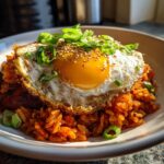 Close-up of vibrant kimchi fried rice topped with a sunny-side-up egg, sesame seeds, and scallions.