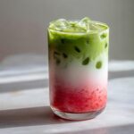 A tall glass showing distinct layers of strawberry puree, milk, and bright green matcha in a refreshing strawberry matcha drink.