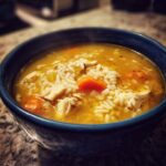 Close-up of a steaming blue bowl filled with Lemon Ginger Turmeric Chicken and Rice Soup, showing rice, shredded chicken, and carrots.