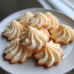 A close-up of several piped, star-shaped butter cookies with golden edges stacked on a white plate.