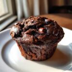 Close-up of a rich, dark chocolate zucchini muffin topped with melted chocolate chips, sitting on a white plate.