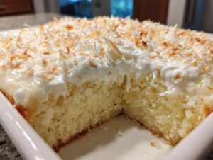 A close-up view of a moist slice cut from a coconut poke cake topped with white frosting and toasted coconut flakes.