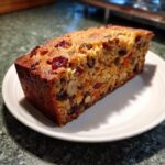 A close-up, appetizing slice of moist fruit cake packed with dried fruit and nuts, served on a white plate.