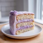 A tall slice of moist lavender cake with three layers of vanilla sponge and purple lavender frosting.
