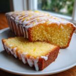 Close-up of a moist lemon loaf cake, sliced, showing a thick white glaze and lemon zest.