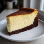 A thick, creamy slice of new york cheesecake featuring a deeply browned top and a dark chocolate crust, served on a white plate.