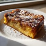 A thick, golden slice of overnight french toast dusted heavily with powdered sugar on a white plate.