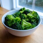 A white bowl filled with vibrant, perfectly steamed broccoli florets resting on a wooden surface near a window.