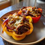 Two baked philly cheesesteak stuffed peppers, one yellow and one red, topped with melted cheese and onions.