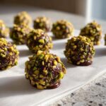 Close-up of rich, dark dubai chocolate balls rolled in chopped green pistachios, sitting on parchment paper.