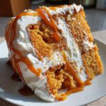 A moist slice of pumpkin poke cake layered with white frosting and drizzled generously with caramel sauce.