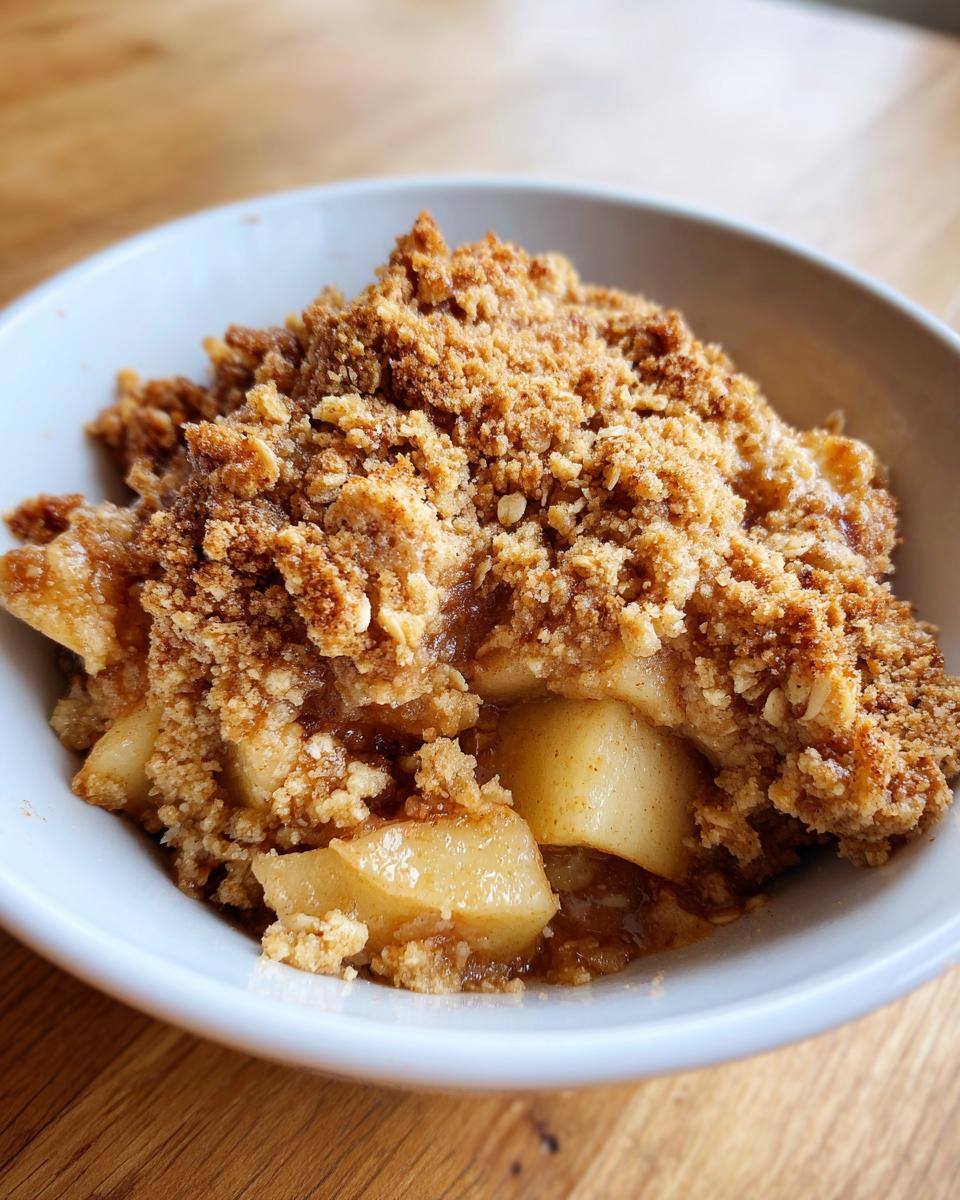 A close-up shot of a warm serving of apple crumble with a thick, golden-brown topping in a white bowl.