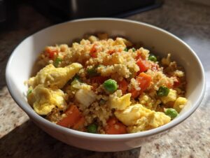 A white bowl filled with vibrant cauliflower rice mixed with scrambled eggs, bright orange carrots, and green peas.