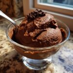 A scoop of rich, dark chocolate mousse topped with shavings, served in a clear glass dish with a spoon.