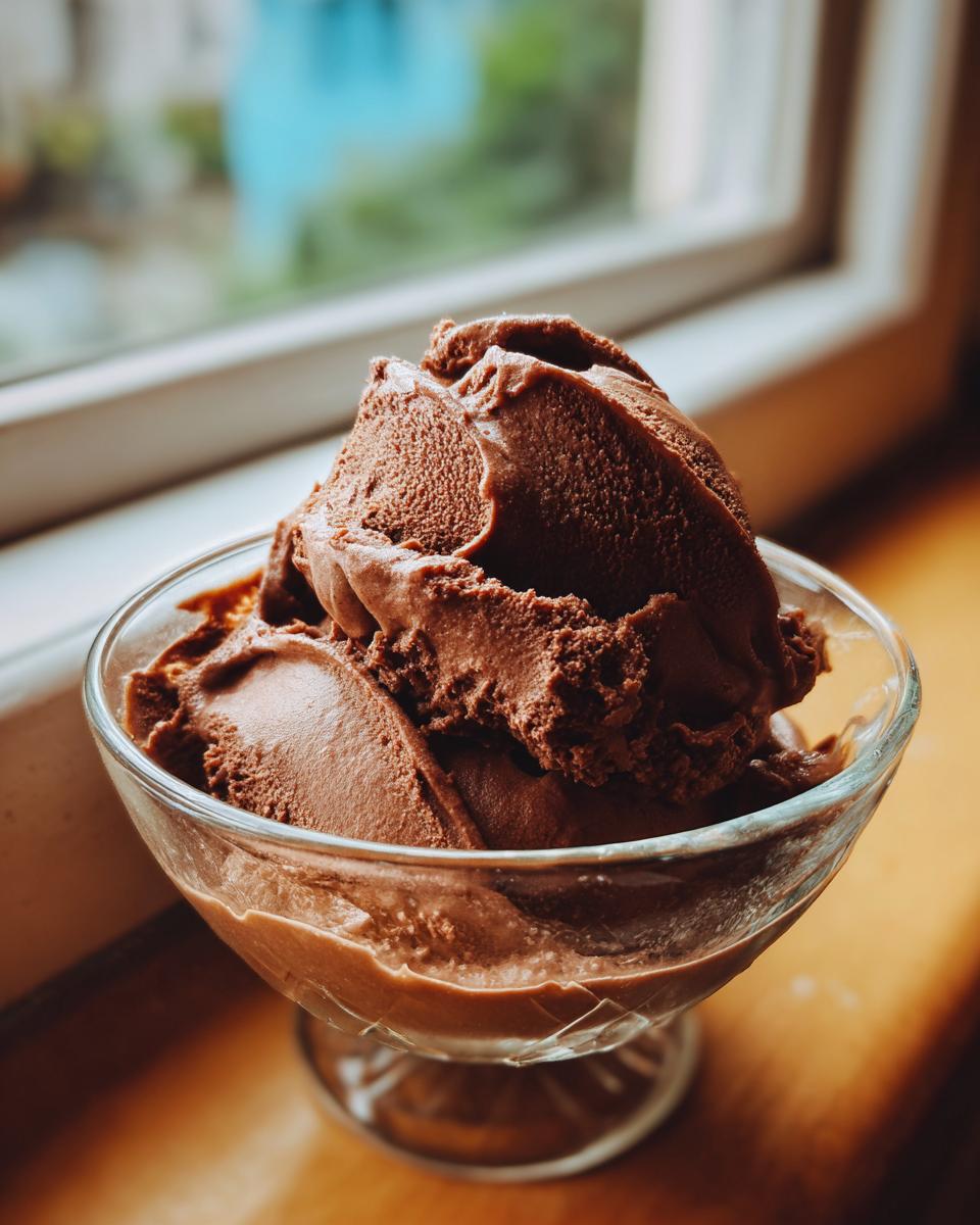 A serving of rich, dark chocolate mousse scooped into a clear glass dessert dish near a window.