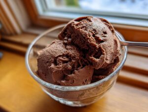 Two scoops of rich, dark chocolate mousse served in a clear glass bowl with a spoon resting inside.