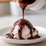 Thick, rich chocolate syrup being poured from a white pitcher onto a scoop of vanilla ice cream.