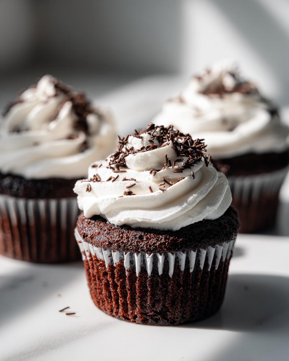 A close-up of a rich, dark chocolate hot chocolate cupcake topped with swirled white frosting and chocolate sprinkles.