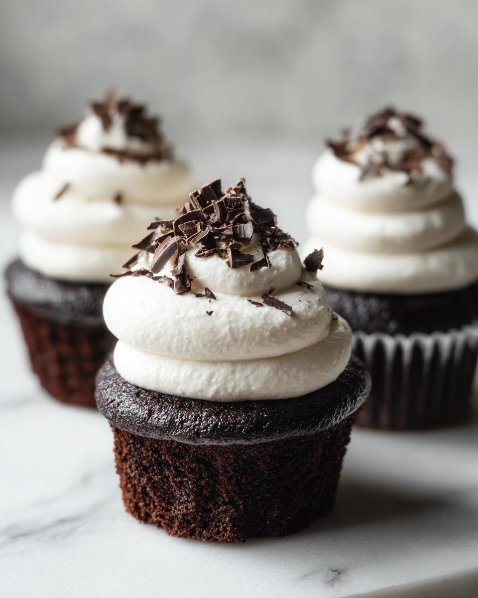Three rich, dark chocolate hot chocolate cupcakes topped with thick white frosting and chocolate shavings.
