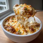 A spoonful of hot, cheesy sausage cream cheese dip is lifted from a white bowl, showing long cheese pulls.