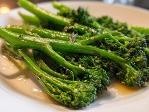 A close-up shot of bright green, glossy sautéed broccolini spears seasoned with garlic on a white plate.