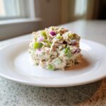 A molded scoop of healthy tuna salad mixed with chopped green celery and purple red onion on a white plate.