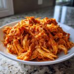 Close-up of shredded buffalo chicken coated in vibrant orange sauce, served in a white bowl.