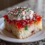 A square slice of moist christmas poke cake topped with red gelatin, whipped cream, and red and green sprinkles.