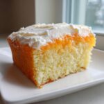 Close-up of a moist slice of Creamsicle Cake showing the yellow sponge, orange glaze, and white frosting.
