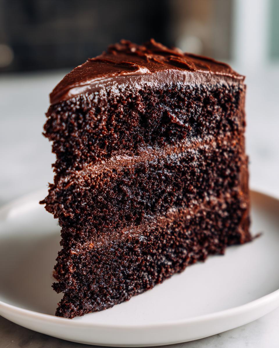 A close-up of a moist, dark slice of homemade chocolate cake layered with rich chocolate frosting.