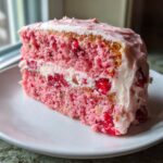 A close-up of a moist slice of pink cherry cake layered with white cream filling and frosted with pink icing.