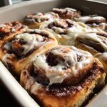 A close-up view of fresh, soft, gooey cinnamon buns baked together in a pan and generously topped with white cream cheese icing.