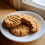 Three soft peanut butter cookies, one broken open, showing classic cross-hatch fork marks on a white plate.