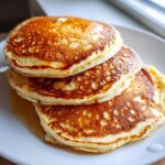 A close-up stack of three golden brown sourdough discard pancakes drizzled with syrup on a white plate.