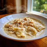 A steaming white bowl filled with creamy chicken and dumplings, topped with shredded chicken and herbs.
