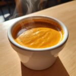 Close-up of a small white cup filled with steaming, rich orange-brown espresso, showing crema texture.