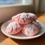 A stack of bright pink, crinkle-topped strawberry cake mix cookies dusted heavily with powdered sugar on a white plate.
