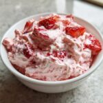 Close-up of a white bowl filled with creamy pink strawberry cheesecake dip topped with fresh strawberry pieces.