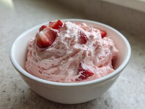 A close-up of fluffy pink strawberry cheesecake dip topped with fresh strawberry slices in a white bowl.