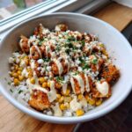A close-up view of a street corn chicken rice bowl topped with seasoned chicken, corn, cotija cheese, and a creamy drizzle.