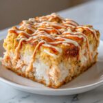 A close-up of a square slice of baked sushi bake topped with drizzles of spicy mayo and white sauce.